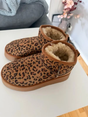 UGG skor  - Säljer ett par snygga leopardmönstrade UGG boots med mjukt beige foder och bruna detaljer. Skorna har en robust sula och är perfekta för att hålla värmen under kalla dagar. De har en låg skaftmodell och är tillverkade i mockaimitation.