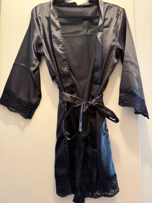 Svart satin morgonrock med spets - Elegant svart morgonrock i glansigt satinmaterial med fina spetsdetaljer vid ärmslut och nederkant. Knytband i midjan och trekvartsärmar ger en lyxig känsla. Perfekt för mysiga hemmakvällar.