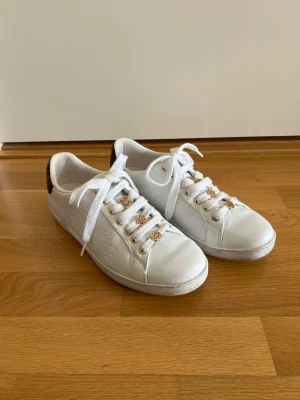 Vita sneakers från Guess med gulddetaljer - Snygga vita sneakers från Guess med guldfärgade detaljer på snörningen och präglade loggor på sidorna. Skorna har vita skosnören och svart detalj med logga på hälen. Tillverkade i konstläder för en clean och fräsch look. De perfekta skorna för sommaren/studenten! 