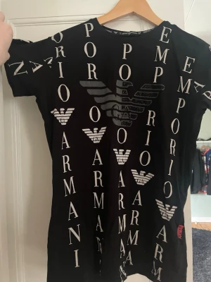 Svart Emporio Armani t-shirt - Snygg svart t-shirt från Emporio Armani med stort logomönster och text tryckt i vitt över hela tröjan. Klassisk rund hals och korta ärmar. Perfekt för dig som gillar streetwear och vill sticka ut med ett exklusivt plagg.