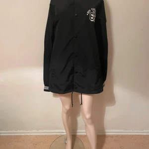 Svart vindjacka från Vans - Snygg svart vindjacka från Vans i storlek medium. Jackan har tryckknappar framtill, vit logga på bröstet och randiga muddar vid ärmsluten. Justerbar dragsko nedtill och klassisk streetstil. Perfekt för dig som gillar enkel och stilren design.