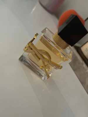 Yves Saint Laurent Libre parfym - Säljer en elegant parfym från Yves Saint Laurent, Libre. Flaskan är genomskinlig med guldiga detaljer och ett svart fyrkantigt lock. Den har en lyxig design med YSL-loggan i metall på framsidan och innehållet är ljust gult.