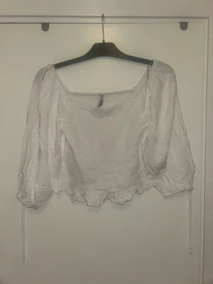 Vit croppad blus med puffärmar - Söt vit croppad blus med puffiga långärmar och snörning vid ärmsluten. Toppen har en fyrkantig ringning och volangkant nedtill. Perfekt för dig som gillar romantisk och trendig stil.