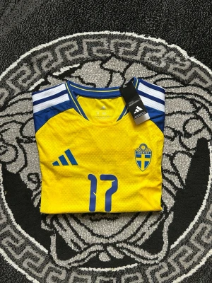 Svensk landslagströja Adidas gul/blå - Säljerw en officiell svensk landslagströja från Adidas i gult och blått med nummer 17 på bröstet. Tröjan har klassiska Adidas-ränder på axlarna, broderad landslagssköld och är tillverkad i ett lätt, ventilerande funktionsmaterial.