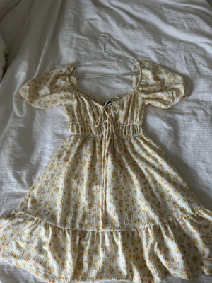 Blommig kortklänning med puffärm - Supersöt kortklänning med små gula och rosa blommor på vit botten. Klänningen har puffärmar, smock i midjan och snörning framtill. Använd på en skolavslutning. 