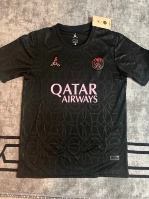 Svart PSG x Jordan matchtröja - Snygg svart PSG matchtröja från Jordan med diskret mönster i tyget. Tröjan har rosa detaljer, PSG-logga och Jordan-logga på bröstet samt Qatar Airways tryckt på magen. Tillverkad i lätt och ventilerande Dri-FIT material, perfekt för träning eller match.
