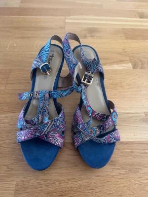 Färgglada mönstrade sandaler med klack - Säljer ett par sandaler från Tamaris med blå klack och färgglada mönstrade remmar i blått, rosa och lila. Remmarna har snygga spännen i guld och korsar över foten för en cool look. Perfekt för dig som vill sticka ut med dina skor!