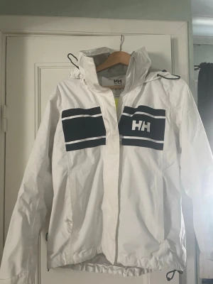 Vit regnjacka från Helly Hansen - Snygg vit regnjacka från Helly Hansen med svarta detaljer och logga på både bröst och rygg. Jackan har huva med dragsko, dragkedja och justerbar nederkant. Perfekt för regniga dagar och outdoor-aktiviteter. Materialet är vattenavvisande och lätt. Defekt bild 3, kanske går bort i tvätt ingen aning då jag inte kunnat bry mig mindre och faktiskt tvätta den