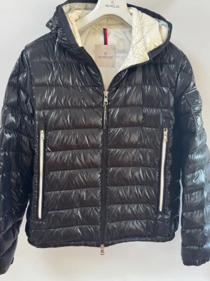 Svart dunjacka Moncler - Snygg svart dunjacka från Moncler med glansig finish och quiltad design. Jackan har två stora dragkedjefickor framtill, huva och vit insida. Perfekt för kalla vinterdagar och riktigt stilren look. Den är självklart äkta och kvitto och påsen till jackan finns här hemma nånstans inget fel skicka 8/10