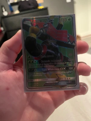 Pokémonkort Tapu Bulu GX - Säljer ett Pokémonkort av typen Tapu Bulu GX med holografisk yta. Kortet har gröna, röda och svarta färger och är inplastat i ett skyddsfodral. Perfekt för samlare eller Pokémonfans.