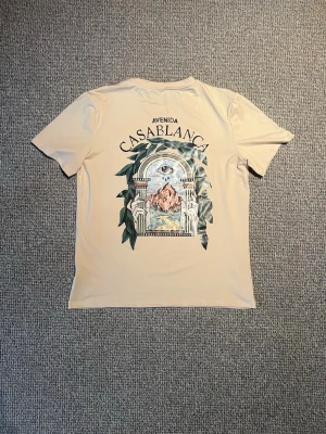 Beige Avenida Casablanca t-shirt - Snygg beige t-shirt från Casablanca med ett stort grafiskt tryck på ryggen som visar ett öga, berg och gröna blad. T-shirten har korta ärmar och en avslappnad passform. Perfekt för dig som gillar unika prints och streetwear vibes.