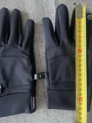 Svarta softshellhandskar LifeHACK - Svarta softshellhandskar från LifeHACK med smidig design och justerbar spänne mellan handskarna. Handskarna har diskret logga på mudden och är perfekta för kyliga dagar. Materialet är vindtätt och stretchigt för extra komfort.