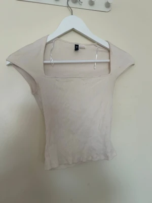 Beige ribbad topp från Divided - Säljer en beige ribbad topp från Divided med fyrkantig urringning och korta ärmar. Toppen är tight i modellen och har en enkel, stilren look som passar perfekt till jeans eller kjol.