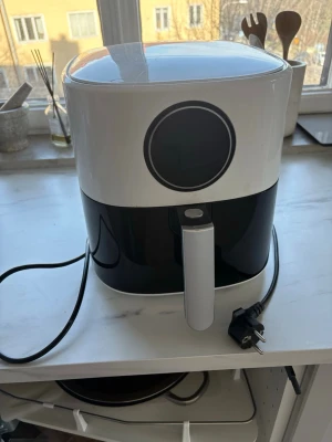 Svart och vit airfryer - Modern airfryer i svart och vit plast med rund form och digital display på framsidan. Utrustad med utdragbar korg och enkel knapp på handtaget. Perfekt för att laga mat snabbt och hälsosamt med minimal olja.