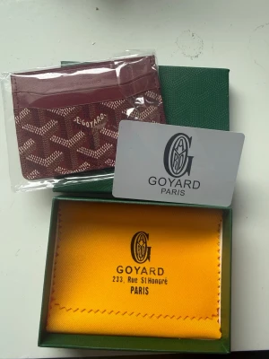 Vinröd korthållare från Goyard - Snyggt vinrött kortfodral från Goyard med klassiskt mönster och logotyp framtill. Fodralet har flera fack för kort och är tillverkat i läder med en elegant och stilren design. Perfekt för dig som vill ha något exklusivt och trendigt.