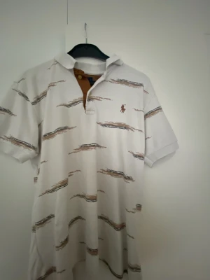Vit mönstrad t-shirt från Polo Ralph Lauren - Vit t-shirt från Polo Ralph Lauren med brunt och svart abstrakt mönster och klassisk logga broderad på bröstet. T-shirten har korta ärmar, krage och knappslå. Perfekt för en avslappnad och stilren look. Krympt lite.