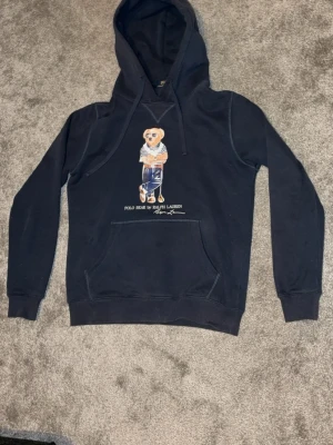 Mörkblå hoodie Polo Bear Ralph Lauren - Mörkblå hoodie från Ralph Lauren med den ikoniska Polo Bear-trycket på bröstet. Klassisk känguruficka framtill och justerbar huva med snören. Mjuk bomullsblandning som känns skön mot huden. Perfekt för en avslappnad och trendig stil.