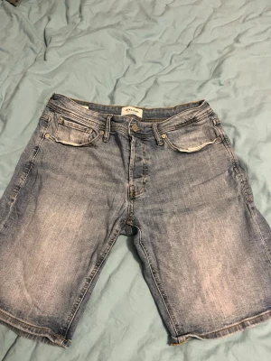 Blå jeansshorts från Jack & Jones - Säljer ett par klassiska blå jeansshorts från Jack & Jones. De har fem fickor, normal passform och snygga slitningar framtill. Perfekta för sommaren och har en avslappnad look med ljus tvätt. Passar till allt och är riktigt bekväma.