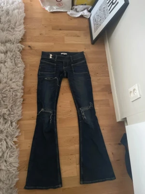 Mörkblå bootcut jeans  - Säljer ett par mörkblå bootcut jeans med slitningar på knäna och detaljerade sömmar. Jeansen har låg midja, fyra fickor och utsvängda ben. 