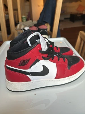 Air Jordan 1 Mid röd/svart/vit Chicago  - Säljer ett par Air Jordan 1 Mid sneakers chicago,  i klassisk röd, svart och vit färgkombination. Skorna har svart swoosh, svarta skosnören och detaljer i läder. Ikonisk Air Jordan-logga på sidan och Jumpman-märke på plösen. Perfekta för dig som gillar streetwear och snygg design.
