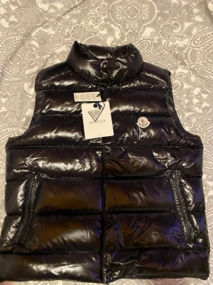 Svart dunväst från Moncler - Snygg svart dunväst från Moncler med glansig finish och hög krage. Västen har två dragkedjeförsedda fickor framtill och klassisk Moncler-logga på bröstet. Tillverkad i ett lätt och värmande material, perfekt för lager-på-lager-stil.