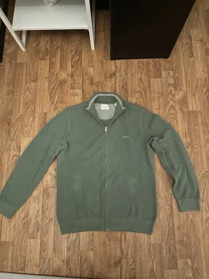 Grön ziptröja från GANT - Snygg grön ziptröja från GANT med diskret logga på bröstet. Tröjan har ståkrage med vita ränder och är ribbstickad vid ärmslut och nederkant. Perfekt för lager på lager och enkel att matcha med jeans eller chinos.