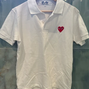 Vit polo t-shirt Comme des Garçons - Vit polo t-shirt från Comme des Garçons Play med klassisk krage och korta ärmar. På bröstet sitter det ikoniska röda hjärtat med ögon. T-shirten är gjord i bomull och har knappslå framtill. Perfekt för dig som gillar stilrena och trendiga plagg.