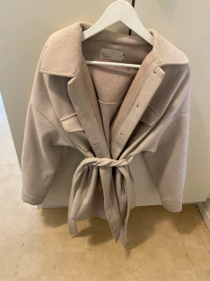 Beige kappa från NLY Trend - Stilren beige kappa från NLY Trend med knytskärp i midjan och bred krage. Kappan har två stora bröstfickor och är tillverkad i ett mjukt, filtliknande material. Perfekt för lager på lager och ger en avslappnad men trendig look. Storlek 34 men passar även 36
