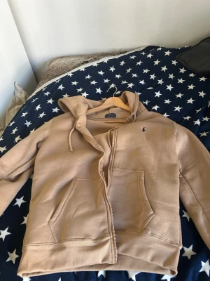 Beige hoodie från Polo Ralph Lauren - Snygg beige hoodie från Polo Ralph Lauren med dragkedja framtill och två stora fickor. Klassisk huva med dragsko och den ikoniska lilla loggan broderad på bröstet. Perfekt för en avslappnad och trendig stil.