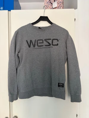 Grå sweatshirt från WESC - Snygg grå sweatshirt från WESC med svart logga framtill. Tröjan har rund halsringning, långa ärmar och ribbade muddar vid ärmslut och nederkant. Perfekt för en avslappnad och stilren look.