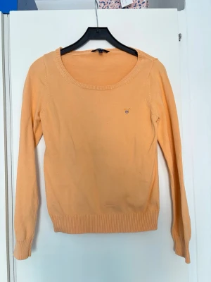 Persikofärgad stickad tröja Gant - Snygg persiko/korallfärgad stickad tröja från Gant med rund halsringning och diskret logga på bröstet. Tröjan har långa ärmar och ribbade muddar vid ärmslut och nederkant. Perfekt för lager-på-lager och enkel att matcha med jeans eller kjol.