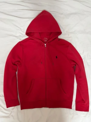 Röd hoodie från Polo Ralph Lauren - Snygg röd hoodie från Polo Ralph Lauren med dragkedja, huva och känguruficka. Klassisk svart logga broderad på bröstet. Mjuk bomullskvalitet och stilren design, perfekt för en avslappnad look.