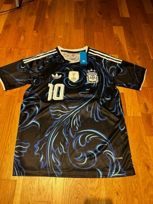 Argentina wc 26 away kit Messi #10 - Snygg svart och blå Argentina fotbollströja från Adidas med Messi och nummer 10 på ryggen. Tröjan har coolt bladmönster, vita detaljer och officiella märken på bröstet. Perfekt för dig som älskar fotboll och vill sticka ut på planen.