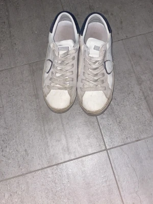 Vita Philippe Model sneakers - Snygga vita sneakers från Philippe Model med marinblå detalj på hälen och klassisk låg siluett. Skorna har vita snören, diskreta sömmar och är tillverkade i läder med mockadetaljer. Perfekta för en clean och stilren look.