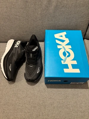 Hoka skor - Säljer ett par svarta Hoka sneakers med vit och svart sula. Skorna har en sportig design med tydlig logga på plösen och sidan. Ovandelen är i mesh för bra andningsförmåga och sulan är tjock för extra komfort. Perfekta för dig som gillar streetwear och sportig stil.