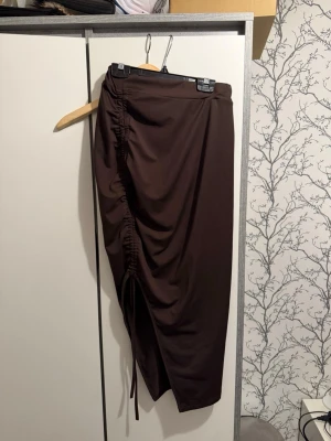Brun långkjol med snöre - Säljer en mörkbrun långkjol med snygg snörningsdetalj på sidan. Kjolen har en draperad effekt och är i ett mjukt, stretchigt material som ger en skön passform. Perfekt för dig som vill ha en trendig och stilren look.