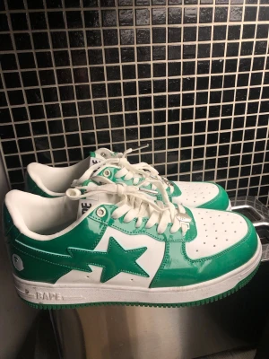 Bape skor - Snygga Bape Sta sneakers i vitt och grönt med glansig finish. Skorna har klassisk låg modell, perforerad tå och ikonisk stjärnlogga på sidan. Vita skosnören med metall-detaljer och BAPE-märke på sulan. Perfekta för dig som gillar streetwear och vill sticka ut.