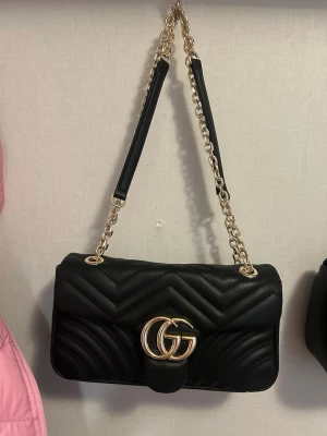 Gucci väska medium marmont  - Säljer en svart quiltad axelväska från Gucci med guldfärgade detaljer och kedjerem. Väskan har GG-logga framtill och ett elegant mönster i materialet. Perfekt storlek för det viktigaste och ger en lyxig vibe till din outfit.
