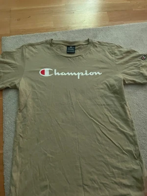Beige Champion t-shirt  - Snygg beige t-shirt från Champion med stor vit logga och röd detalj på bröstet. Klassisk rund halsringning och korta ärmar. Liten Champion-logga även på ärmen. Perfekt till en avslappnad streetwear-look.