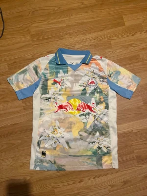 FC Salzburg x Puma matchtröja - Unik FC Salzburg matchtröja från Puma med färgstarkt blommönster i blått, gult, grönt och orange. Tröjan har klassisk krage med blå och gula detaljer, Red Bull-logga på bröstet och klubbmärke. Perfekt för dig som vill sticka ut på läktaren eller träningen.