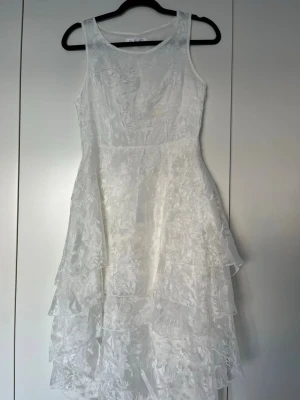 Vit ärmlös klänning med volanger - Snygg vit klänning med ärmlös design och flera lager volanger i kjolen. Klänningen har en lätt glansig yta och subtilt blommönster i tyget. Perfekt för dig som vill ha en romantisk och stilren look.