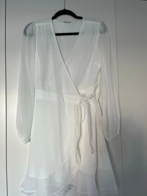 Vit omlottklänning med volang - Säljer en vit omlottklänning med långa, transparenta ärmar och knytband i midjan. Klänningen har volangdetaljer nertill och är gjord i ett lätt, luftigt material som ger en romantisk känsla. Perfekt för sommarens alla tillfällen.