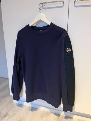 Colmar tröja - Mörkblå stickad tröja från Colmar med quiltad detalj längs ena ärmen och rund halsringning. Tröjan har ribbade muddar vid ärmslut och nederkant samt en Colmar-logga på ärmen. Perfekt för dig som gillar stilrena och sportiga plagg.