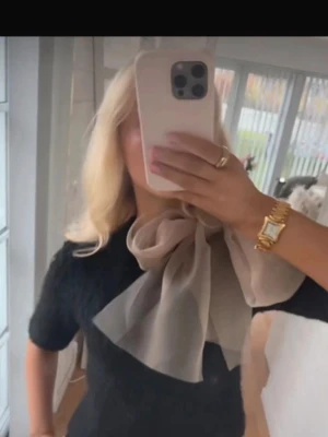 Svart stickad tröja med beige rosett - Supersöt svart stickad tröja med korta ärmar och en stor beige rosett i tunn chiffong vid halsen. Tröjan har en fluffig struktur och ger en trendig look som sticker ut. Perfekt för dig som vill ha något unikt och stilrent i garderoben.
