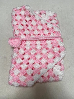 Handvirkat bokfodral rosa/vit - Unikt bokfodral som är handvirkat i mjukt garn med rutmönster i rosa och vitt. Fodralet har ett snöre med ett litet hjärta som dekoration och stängning. Perfekt för att skydda böcker (ca 13-16cm x 19-22cm) eller anteckningsböcker och ger en personlig touch till din läsning.