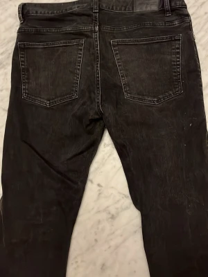 Svarta raka jeans - Säljer ett par svarta jeans med klassisk femficksmodell och raka ben. Jeansen har bälteshällor, silverfärgade nitar och knapp, samt tydliga sömmar. Materialet är denim i bomull. Perfekta för en avslappnad och stilren look.