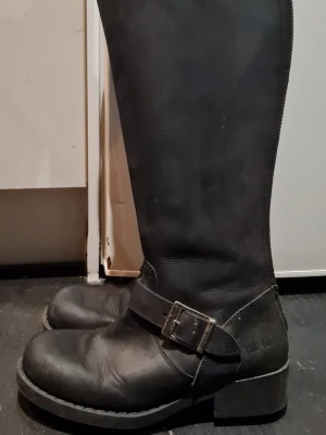 Svarta läderstövlar med spänne - Säljer ett par svarta höga läderstövlar från Johnny Bulls med grov sula, dekorativt spänne och dragkedja längs baksidan. Klassisk bikerstil som passar perfekt till jeans eller kjol. Stövlarna har rund tå och robust klack.