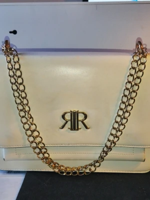 White handbag from River Island - Stylish white handbag from River Island with a gold-colored chain strap and logo on the front. The bag has a stylish, square shape and gold details. The material is smooth artificial leather, croc embossed on the sides. Perfect for those who like trendy accessories.