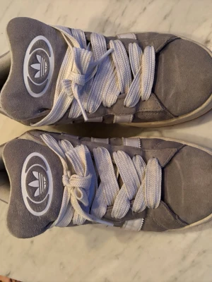 Grå Adidas sneakers med vita ränder - Säljer ett par grå sneakers från Adidas med klassiska vita ränder på sidan och stor Adidas-logga på plösen. Skorna har vita snören och en sula i gummi med beige kant. Ovandelen är i mockaliknande material och modellen är retroinspirerad. Använda ett fåtal gånger 