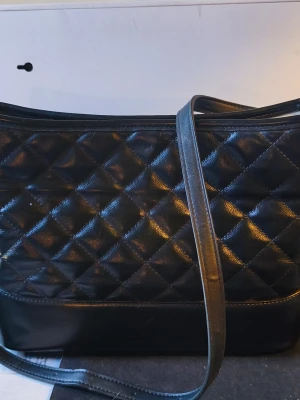 Black quilted handbag  - Stylish black shoulder bag with quilted pattern and long shoulder strap. The bag is made of glossy leather-like material and has silver-colored metal details. Perfect for those who like classic and stylish design.